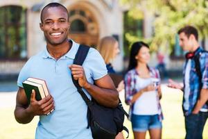 das Unileben genießen. hübscher junger afrikanischer mann, der bücher hält und lächelt, während er mit seinen freunden, die im hintergrund plaudern, gegen die universität steht foto