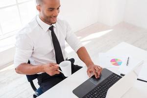 mit Freude arbeiten. Draufsicht eines selbstbewussten jungen afrikanischen Mannes im Hemd, der am Laptop arbeitet und lächelt, während er an seinem Arbeitsplatz sitzt foto