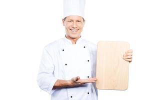 Sonderangebot vom Küchenchef. Selbstbewusster reifer Koch in weißer Uniform, der ein Holzschneidebrett hält und es mit einem Lächeln zeigt, während er vor weißem Hintergrund steht foto