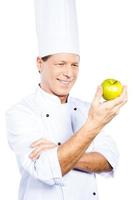 Kochen Sie nur gesundes Essen. Selbstbewusster, reifer Koch in weißer Uniform, der grünen Apfel hält und ihn mit einem Lächeln betrachtet, während er vor weißem Hintergrund steht foto