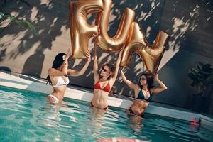Attraktive junge Frauen in Badebekleidung lächeln und heben Luftballons hoch, während sie draußen im Pool stehen foto