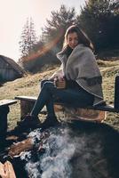 Aufwärmen. Schöne junge Frau, bedeckt mit einer Decke, die einen Becher hält, während sie auf der Bank am Lagerfeuer sitzt foto