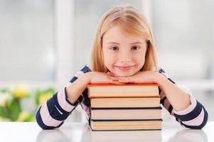 Ich liebe es, ein fröhliches kleines Mädchen zu studieren, das sich am Bücherstapel lehnt, während es am Tisch sitzt foto