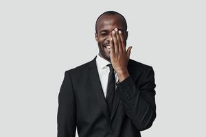 Verspielter junger afrikanischer Mann in Abendkleidung, der die Hälfte seines Gesichts mit der Hand bedeckt und lächelt, während er vor grauem Hintergrund steht foto