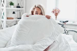 Montagmorgen. Attraktive junge Frau, die die Hälfte ihres Gesichts mit einer Decke bedeckt und in die Kamera schaut, während sie zu Hause im Bett sitzt foto