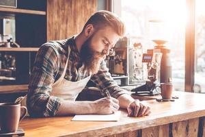 neue Kaffeerezepte aufschreiben. Junger bärtiger Mann in Schürze schreibt in Notizbuch, während er sich im Café an die Theke lehnt foto