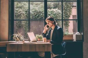 Arbeiten vom Café. Fröhlicher junger Mann in eleganter Freizeitkleidung, der auf den Laptop schaut und mit dem Handy telefoniert, während er in der Nähe des Fensters im Kreativbüro oder Café sitzt foto