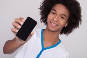 zeigt sein neues Handy. Fröhlicher afrikanischer Teenager, der sein Handy zeigt und lächelt, während er isoliert auf grauem Hintergrund steht foto