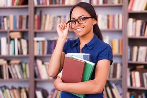 sich sicher fühlen, wenn es um Prüfungen geht. Fröhliche afrikanische Studentin, die Bücher hält und lächelt, während sie in der Bibliothek steht foto