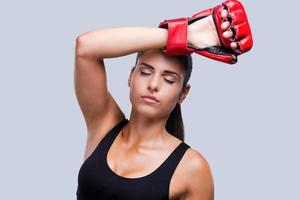 Müdigkeit nach dem Training. Attraktive junge sportliche Frau in Boxhandschuhen, die ihre Stirn berührt und die Augen geschlossen hält, während sie vor grauem Hintergrund steht foto