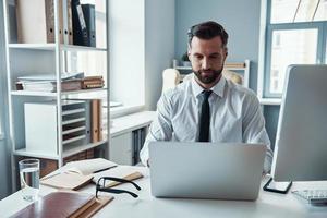 Konzentrierter junger Mann in Hemd und Krawatte, der mit dem Computer arbeitet, während er im Büro sitzt foto