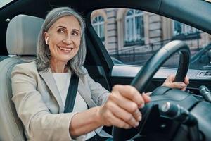 Reife schöne Frau in eleganter Freizeitkleidung, die lächelt und fährt, während sie auf dem Vordersitz des Autos sitzt foto