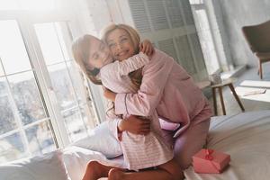 glücklich, zusammen zu sein. süßes kleines mädchen, das ihre mutter umarmt, während sie zu hause auf dem bett sitzt foto