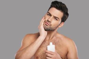 sah früher perfekt aus. hübscher junger Mann, der Aftershave-Lotion aufträgt und lächelt, während er vor grauem Hintergrund steht foto