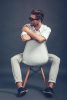 Herr Stil. voller Länge von selbstbewusstem jungen gutaussehenden Mann mit Sonnenbrille, der wegschaut, während er auf einem Stuhl vor grauem Hintergrund sitzt foto