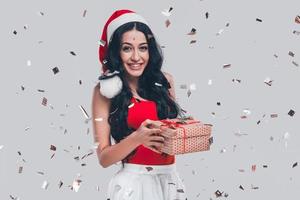 Frohe Weihnachten Attraktive junge Frau mit Weihnachtsmütze, die eine Geschenkbox hält und lächelt, während sie vor grauem Hintergrund mit Konfetti um sie herum steht foto