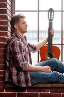 junger und kreativer Gitarrist. Seitenansicht eines hübschen jungen Mannes, der eine Akustikgitarre hält, während er auf der Fensterbank sitzt foto