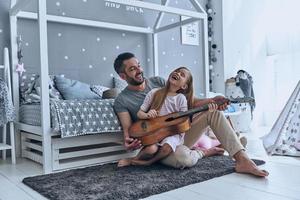 Musik macht so viel Spaß. Der junge Vater bringt seiner kleinen Tochter das Gitarrespielen bei und lächelt, während er im Schlafzimmer auf dem Boden sitzt foto