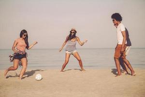 aktive Spiele mit Freunden. gruppe fröhlicher junger leute, die mit fußball am strand mit meer im hintergrund spielen foto
