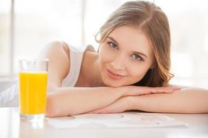 sich frisch und gesund fühlen. Schöne junge Frau, die in die Kamera schaut und lächelt, während sie mit einem Glas Orangensaft am Tisch sitzt foto