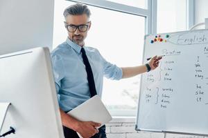 männlicher englischlehrer, der whiteboard zeigt, während er webkonferenz im klassenzimmer hat foto