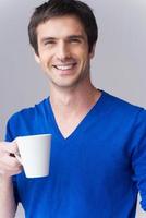heißes Getränk genießen. hübscher junger Mann im blauen Pullover, der Kaffeetasse hält und lächelt, während er vor grauem Hintergrund steht foto
