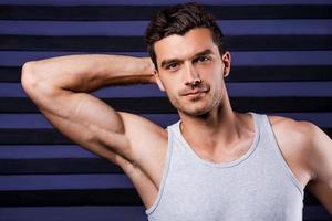 überzeugt von seinem perfekten Körper. sportlicher junger Mann im Tanktop, der die Hand hinter dem Kopf hält und in die Kamera blickt, während er vor gestreiftem Hintergrund steht foto