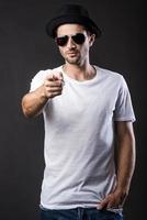 cool und trendy. hübscher junger Mann mit Sonnenbrille und Fedora, der die Hände in den Taschen hält, während er vor schwarzem Hintergrund steht foto