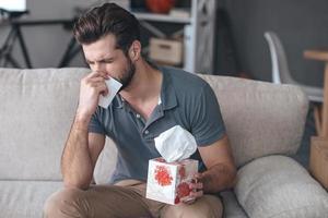 schreckliche allergie. frustrierter gutaussehender junger mann, der niest und taschentuch benutzt, während er zu hause auf der couch sitzt foto