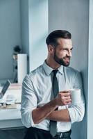 hübscher junger mann in hemd und krawatte, der wegschaut und eine tasse hält, während er im büro steht foto