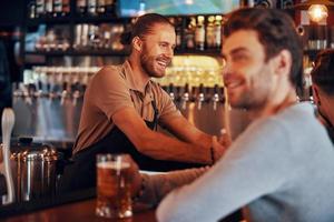 Fröhlicher Barkeeper, der Getränke serviert, während der junge lächelnde Mann im Pub Bier im Vordergrund genießt foto