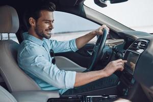 genießt seine Fahrt. hübscher junger Mann in eleganter Freizeitkleidung, der lächelt, während er ein Statusauto fährt foto