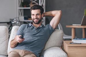 tolle tv-show. hübscher fröhlicher junger mann, der fernbedienung hält und fernsieht, während er zu hause auf der couch sitzt foto