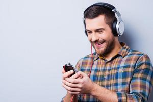 liebe diese Musik. hübscher junger Mann mit Kopfhörern, der Handy hält und lächelt, während er vor grauem Hintergrund steht foto
