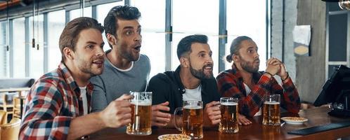 Jubelnde junge Männer in Freizeitkleidung, die Bier trinken und Sportspiele beobachten, während sie in der Kneipe sitzen foto