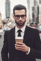 Kaffee unterwegs genießen. selbstbewusster junger Mann im vollen Anzug mit Kaffeetasse und Blick in die Kamera, während er im Freien mit Stadtbild im Hintergrund steht foto