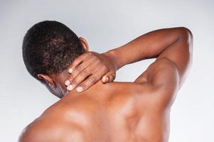 Nackenschmerzen. Rückansicht eines jungen afrikanischen Mannes ohne Hemd, der seinen Hals berührt, während er vor grauem Hintergrund steht foto