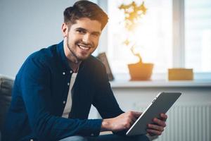 Dieses Tablet übertrifft seine Erwartungen. Seitenansicht eines hübschen jungen Mannes, der sein digitales Tablet benutzt und mit einem Lächeln in die Kamera schaut, während er zu Hause auf der Couch sitzt foto