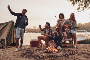 gruppe junger leute in freizeitkleidung, die selfie machen und lächeln, während sie eine strandparty am lagerfeuer genießen foto