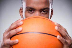 fertig zu spielen. Nahaufnahme eines jungen, hemdlosen afrikanischen Mannes, der einen Teil seines Gesichts hinter einem Basketballball versteckt und in die Kamera blickt, während er vor grauem Hintergrund steht foto