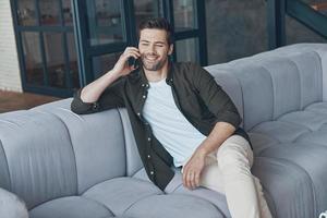 hübscher junger mann, der auf dem smartphone spricht und wegschaut, während er zu hause auf dem sofa sitzt foto