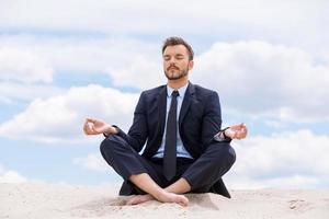 Ruhe bewahren in seiner Seele. hübscher junger Geschäftsmann, der beim Sitzen im Lotussitz auf Sand und gegen blauen Himmel meditiert foto