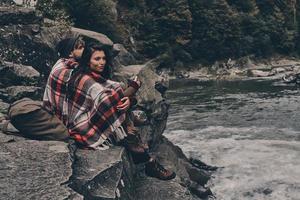 dem Gesang der Natur lauschen. Schönes junges Paar, das mit einer Decke bedeckt ist und wegschaut, während es auf den Felsen in der Nähe des Flusses sitzt foto