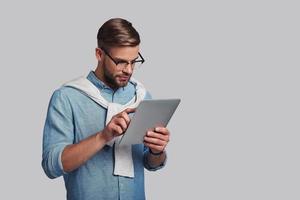 untersucht sein digitales Tablet. ernsthafter junger Mann mit Brille, der mit einem digitalen Tablet arbeitet, während er vor grauem Hintergrund steht foto