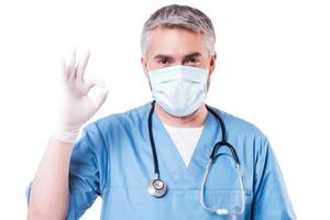 Chirurg gestikuliert ok. reifer Arzt mit grauen Haaren in OP-Maske und Handschuhen, die ein Ok-Zeichen gestikulieren, während sie isoliert auf Weiß stehen foto