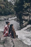 gemeinsam wohlfühlen. Schönes junges Paar, das mit einer Decke bedeckt ist und wegschaut, während es auf den Felsen in der Nähe des Flusses sitzt foto