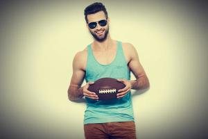 will mit mir spielen selbstbewusster junger gutaussehender mann mit sonnenbrille, der american-football-ball hält und lächelt, während er vor weißem hintergrund steht foto