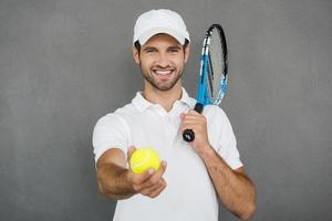 Begleiten Sie mich hübscher junger Mann in Sportkleidung, der Tennisschläger auf seiner Schulter trägt und Tennisball ausstreckt, während er vor grauem Hintergrund steht foto