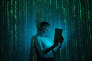 Selbstbewusste Frau in futuristischer Brille mit digitalem Tablet vor dunklem Hintergrund mit grünen Symbolen foto