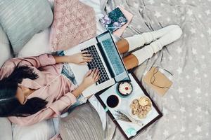 Blick von oben auf die schöne junge Frau im Schlafanzug mit Laptop, während sie sich zu Hause im Bett ausruht foto